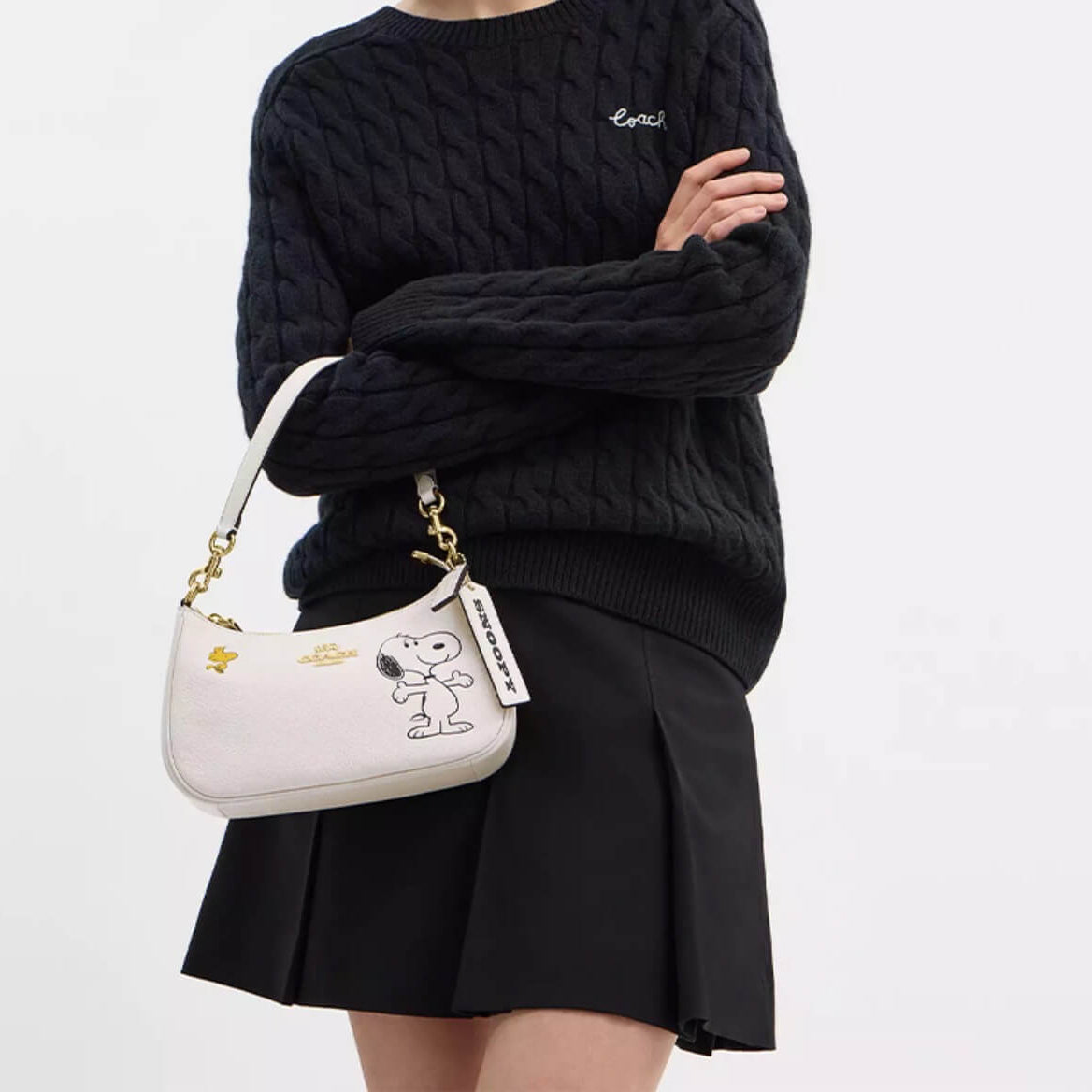 A model wearing a black cable-knit sweater and black pleated skirt carries a white leather shoulder bag featuring a cartoon character graphic and gold hardware.