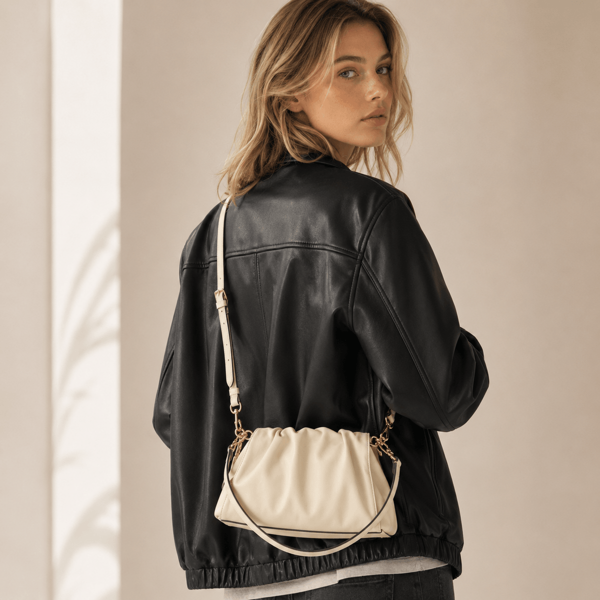 A woman wearing a black faux-leather jacket and dark jeans, shown from the back and side, carrying a cream-colored faux-leather shoulder bag with gold-tone hardware.