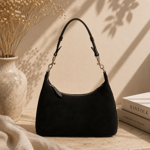 A black suede shoulder bag with a curved top and a single shoulder strap, styled on a tabletop next to a beige ceramic vase with dried flowers and a stack of books.