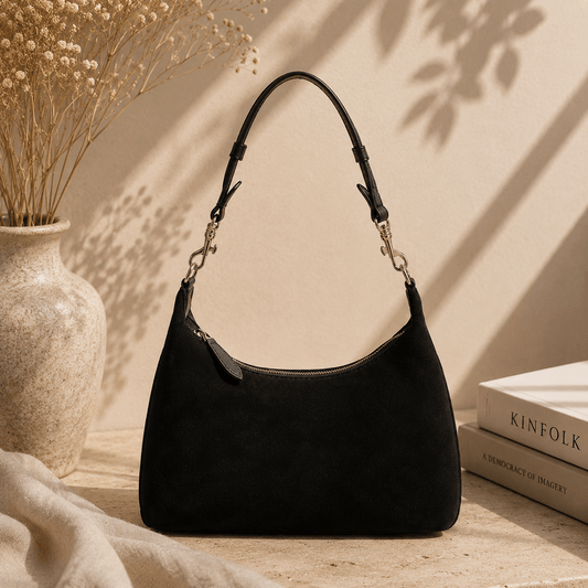 A black suede shoulder bag with a curved top and a single shoulder strap, styled on a tabletop next to a beige ceramic vase with dried flowers and a stack of books.