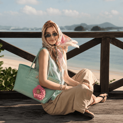 A woman sits on a wooden deck overlooking a beach, wearing a mint green sleeveless top, beige pants, and a pink patterned headscarf. She is carrying a large mint green tote bag with a pink crocheted flower accent.