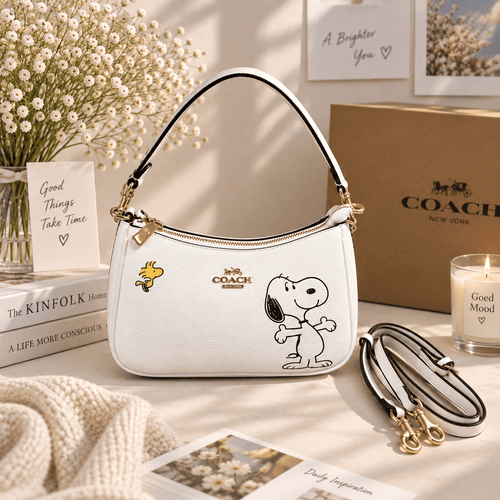 A white  handbag with a black and white Snoopy print, displayed in a styled setting with a Coach box, books, flowers, and candles.
