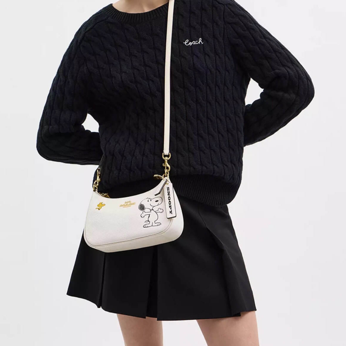 A model wearing a black cable-knit sweater and black pleated skirt, carrying a white crescent-shaped shoulder bag with a cartoon character print and gold hardware.