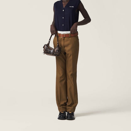 A model is shown from the chest down wearing brown wide-leg trousers, a navy sleeveless button-up shirt, and black loafers, holding a small brown handbag against a plain beige studio background.