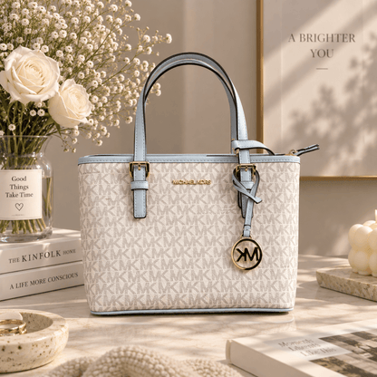 A Michael Kors tote bag in white micro-printed leather with light blue handles and trim, styled on a tabletop with books, a vase of roses, and a framed print in a warm, sunlit interior.