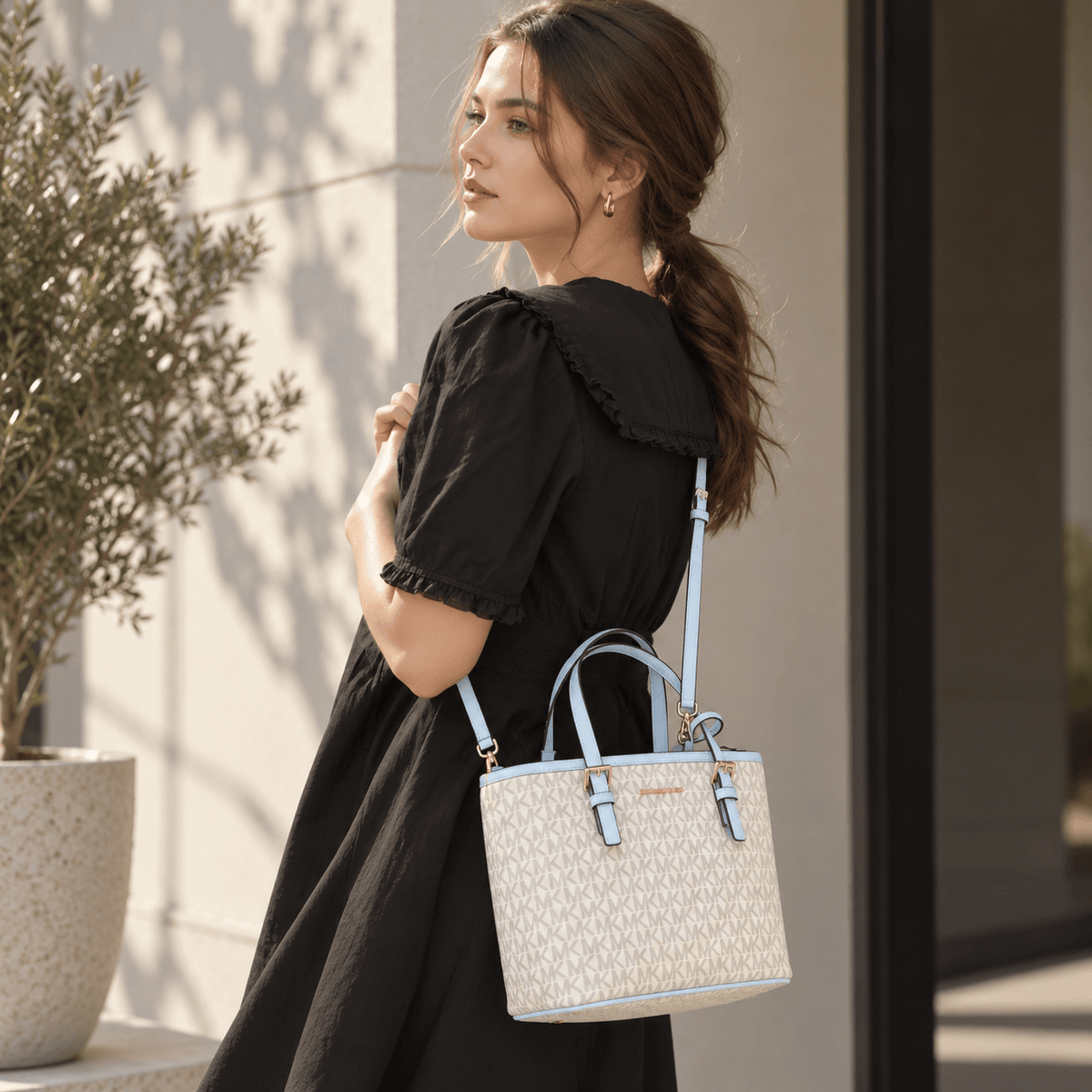 A woman in a black dress carries a structured tote bag with a cream and light blue monogram pattern, light blue handles, and a light blue shoulder strap in an outdoor setting.