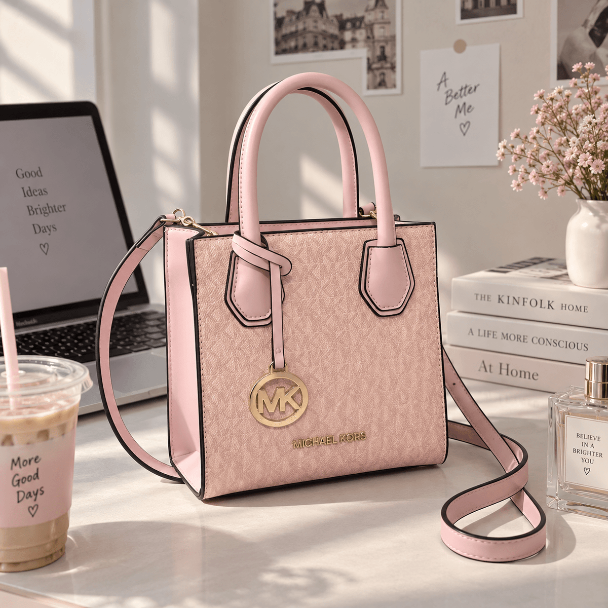 A pink Michael Kors handbag with a monogram pattern, gold-tone hardware, and a matching shoulder strap, styled on a desk with a laptop, books, flowers, and a drink.