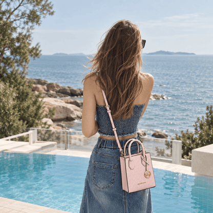A woman stands by a pool overlooking the sea, wearing a pink structured handbag with a long strap and gold-tone charm. The scene is outdoors with rocks, trees, and blue water in the background.