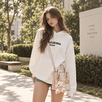 A woman stands outdoors on a sunny paved walkway wearing a white oversized hoodie with black text reading 'BLOOMING BRIGHT' and carrying a small pink and beige patterned shoulder bag with a teddy bear charm.