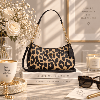 A Coach shoulder bag with a beige and black leopard print pattern, black leather trim, and gold-tone chain handles, styled on a marble tabletop with books, flowers, sunglasses, and a coffee cup.