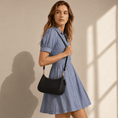 A woman wearing a light blue chambray short-sleeve dress and carrying a black shoulder bag with gold-tone hardware, standing against a warm beige wall with soft window shadows.