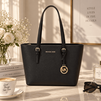 A black Michael Kors tote bag with gold-tone hardware, displayed on a marble tabletop with a coffee cup, sunglasses, flowers, and framed wall art in a styled interior setting.